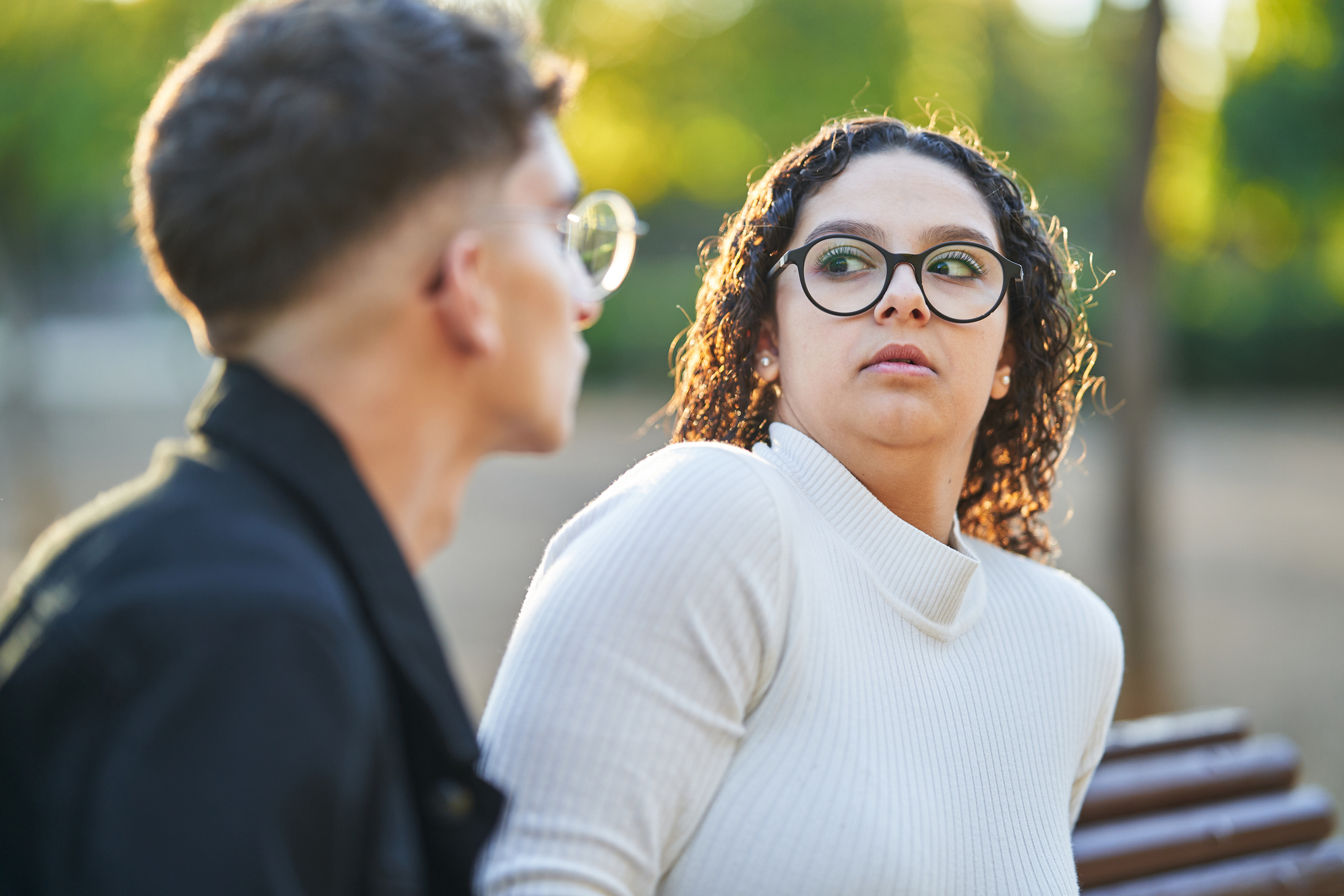 Two people sitting on a bench outdoors, a woman in glasses looks at a man with a curious expression