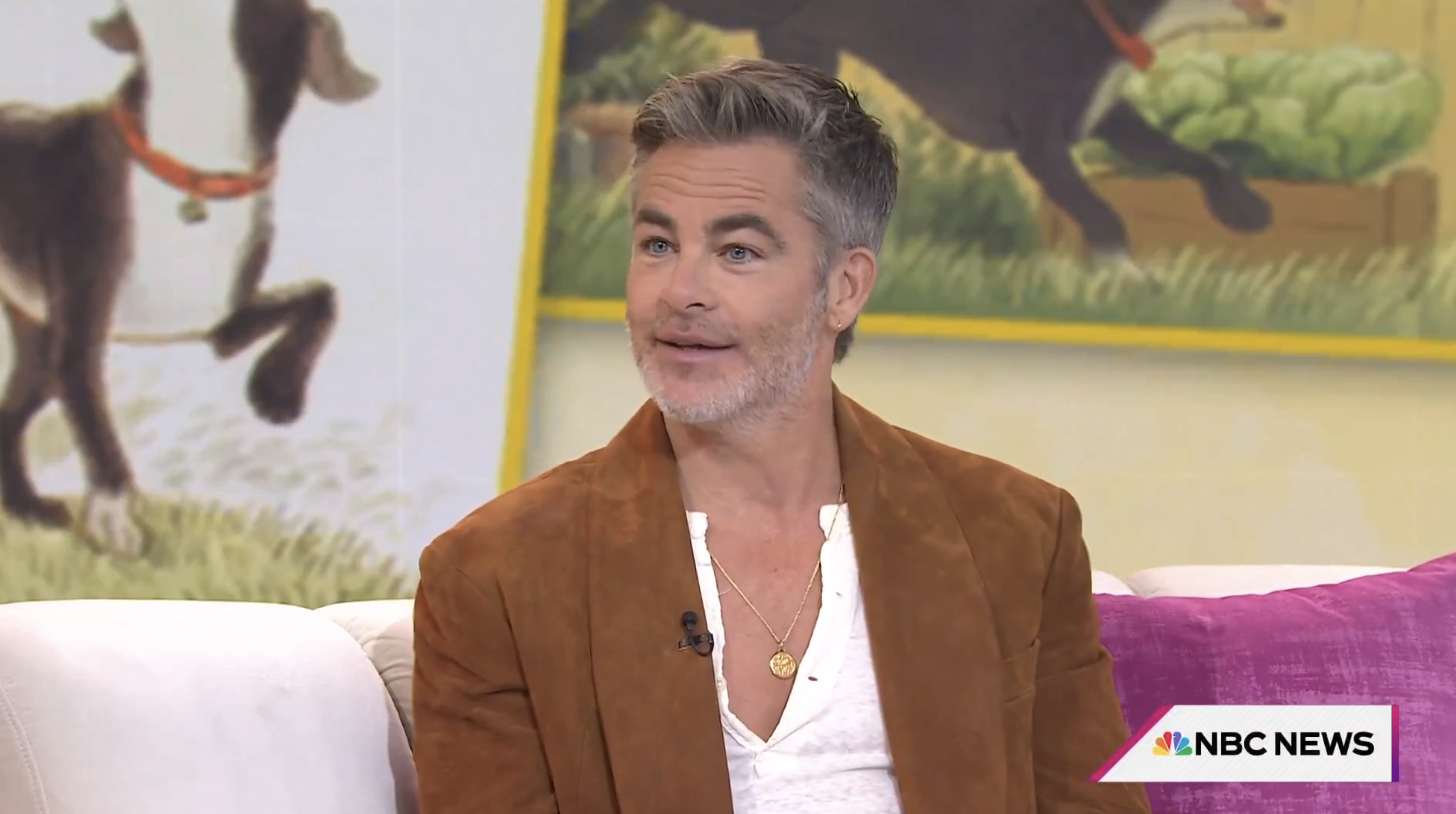 Chris Pine wearing a suede jacket over a shirt, sitting on a couch with a colorful backdrop