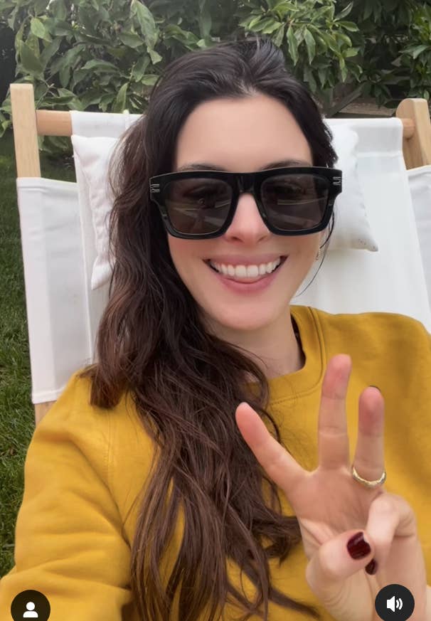 Anne Hathaway with long hair and sunglasses, wearing a sweater, smiles while holding up three fingers, sitting outdoors