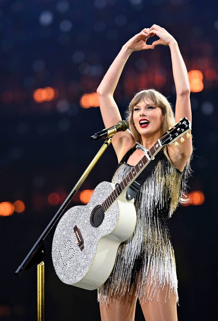 The person is performing on stage, wearing a shimmering fringe dress, holding a glittery guitar, and forming a heart with their hands