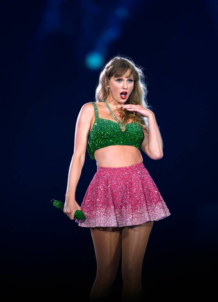 A person wearing a sparkly green top and pink skirt performs on stage with a green microphone