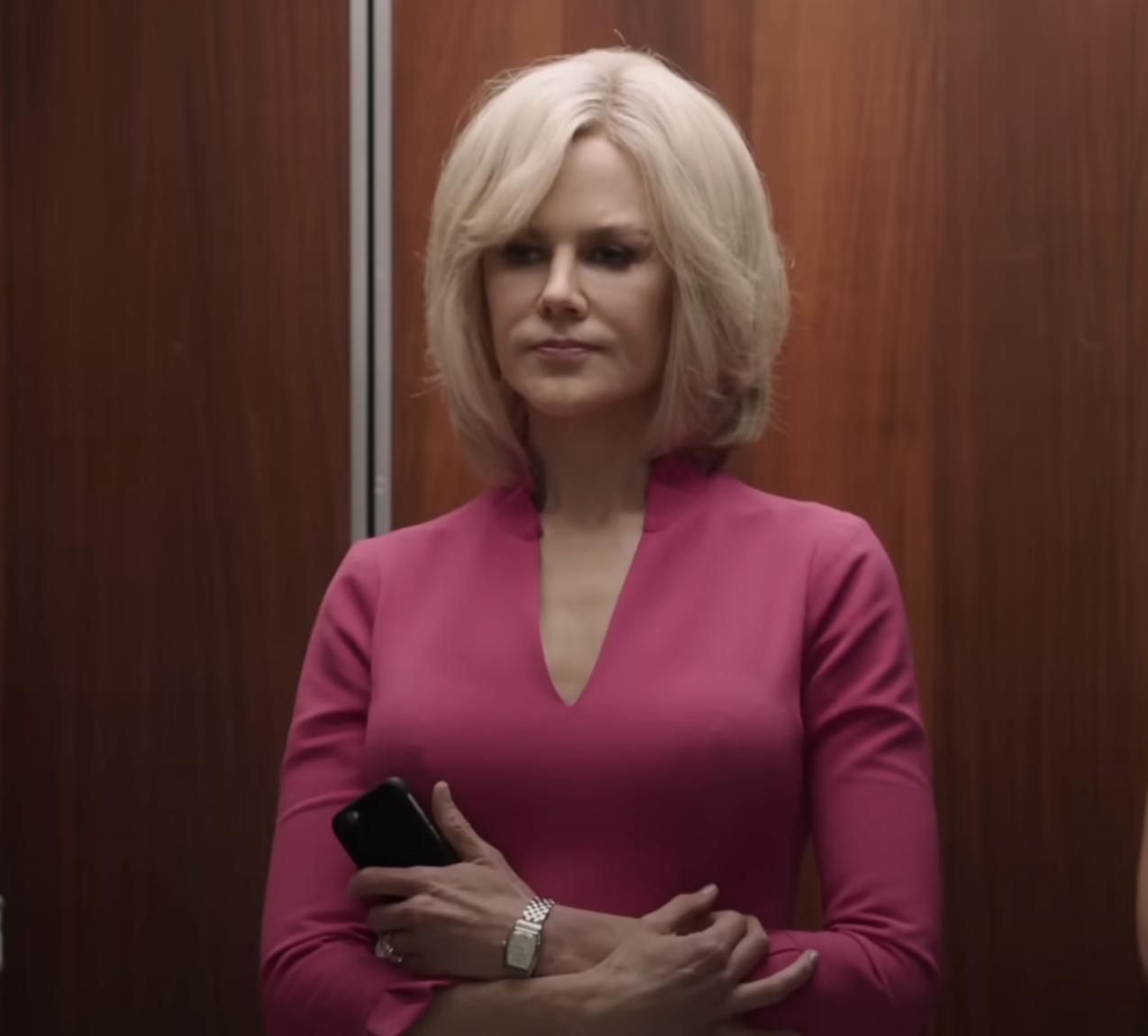 A woman with short, styled hair stands in an elevator holding a phone, wearing a fitted, sophisticated outfit
