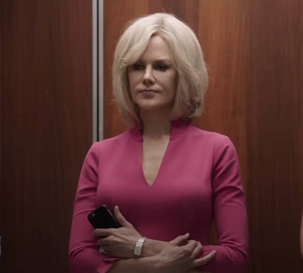 A woman with short, styled hair stands in an elevator holding a phone, wearing a fitted, sophisticated outfit