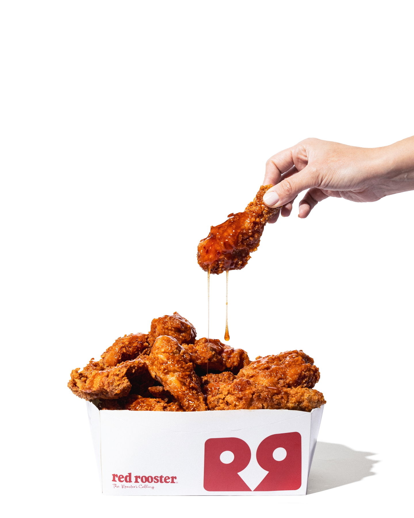 A hand holding a piece of fried chicken above a box labeled &quot;Red Rooster&quot; filled with more fried chicken; sauce drips from the chicken