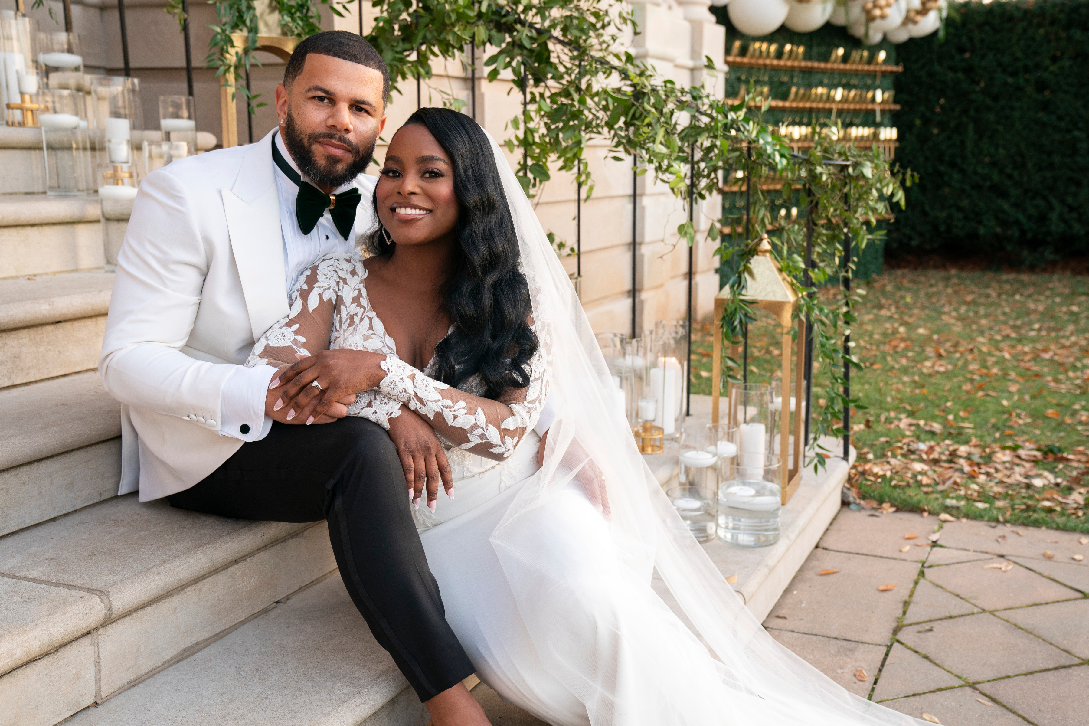 A couple poses on steps in wedding attire; the woman in a gown with lace sleeves, the man in a white tuxedo jacket and black pants