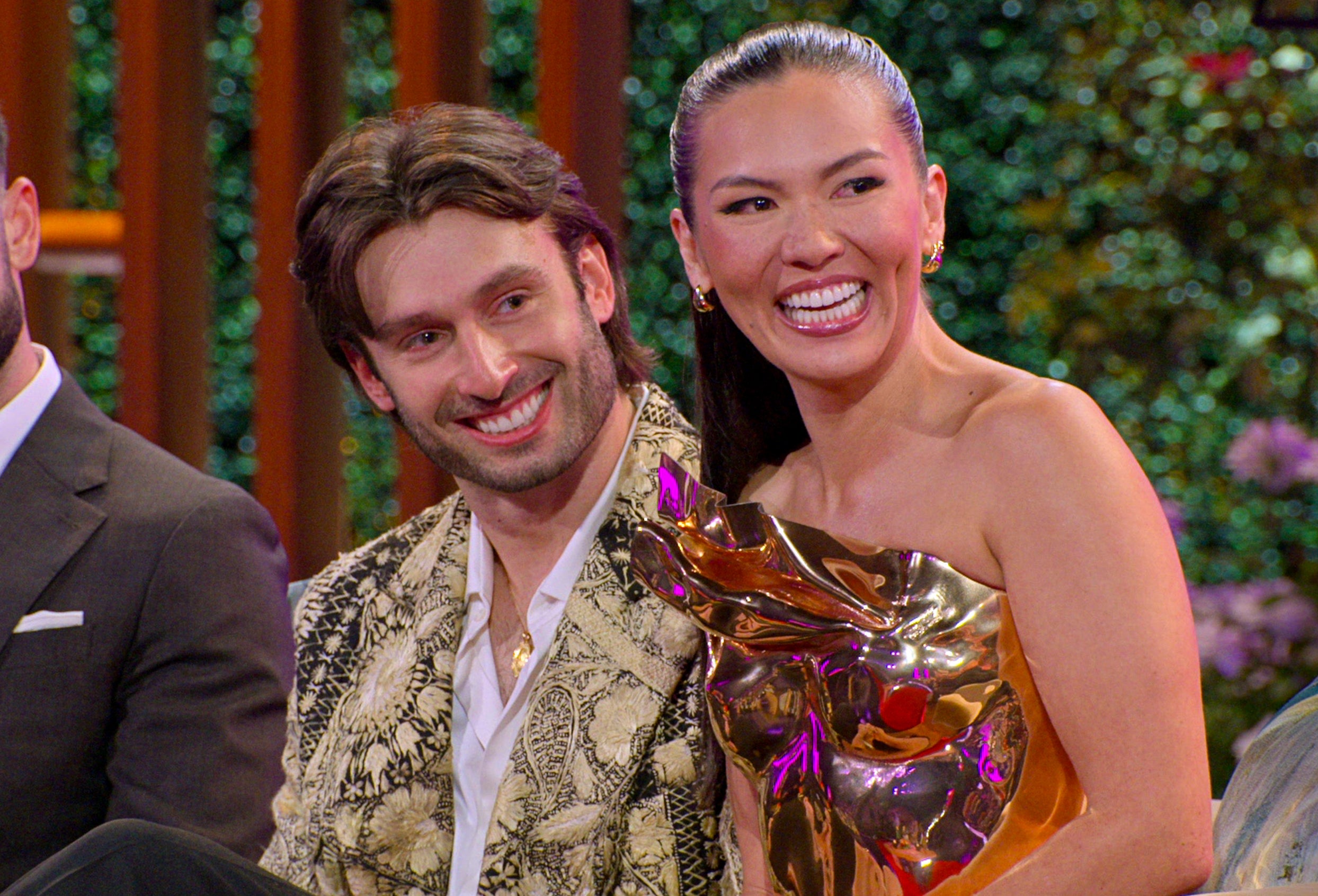 A couple smiles at a TV event; the woman wears an elegant, reflective gold dress, the man is in a patterned jacket