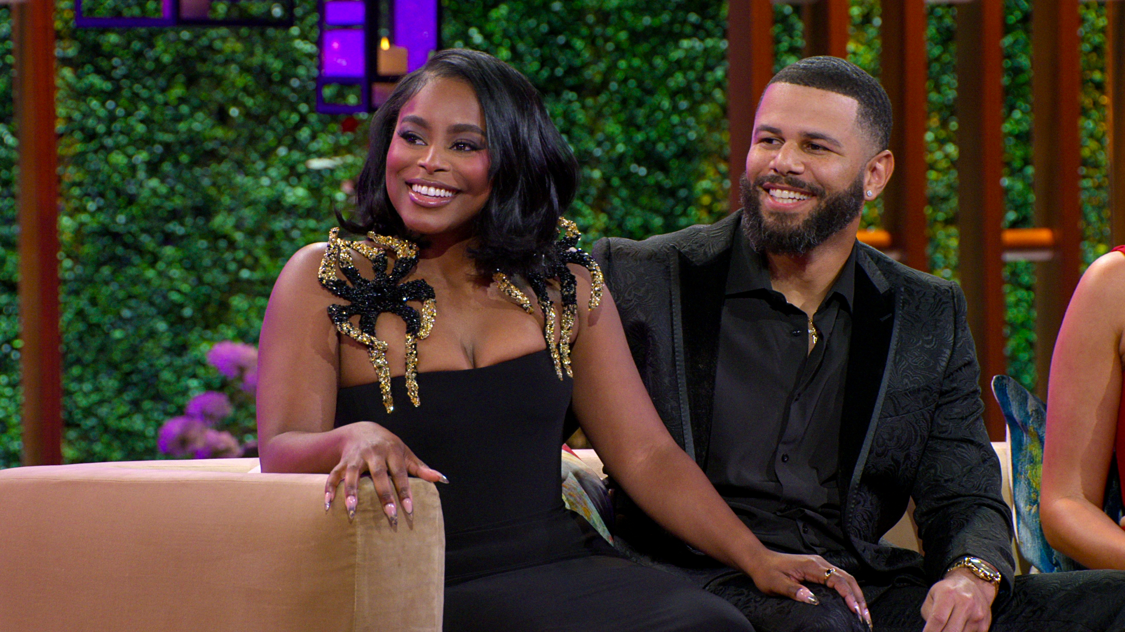 Two individuals sit on a couch during a talk show appearance; the woman wears a black dress with decorative shoulders