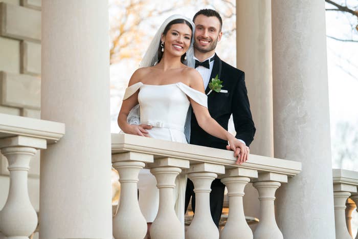 A bride in an elegant off-shoulder gown and a groom in a classic tuxedo pose on a balcony