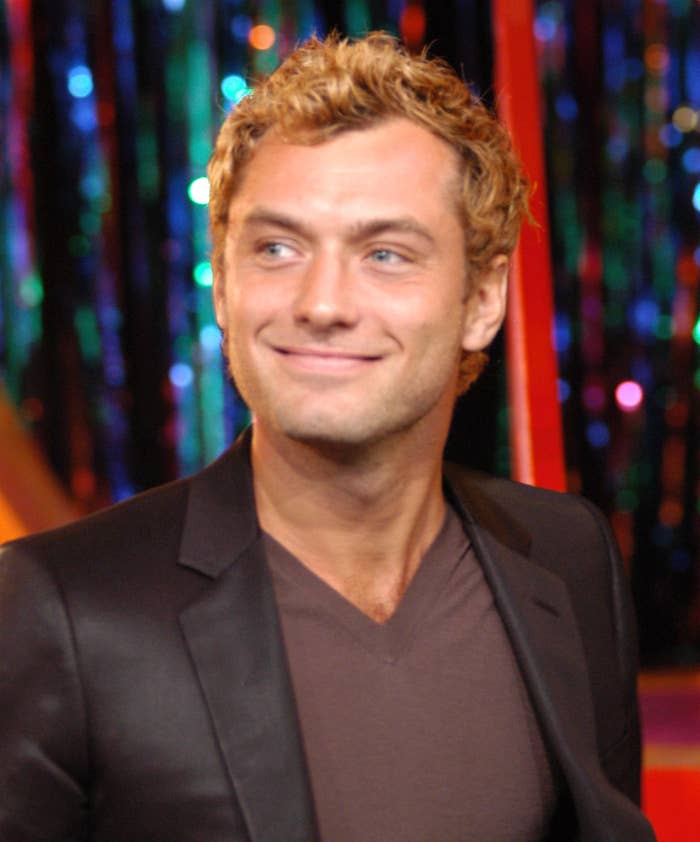 Jude Law wearing a dark blazer over a V-neck shirt, smiling, standing against a colorful, sparkly backdrop