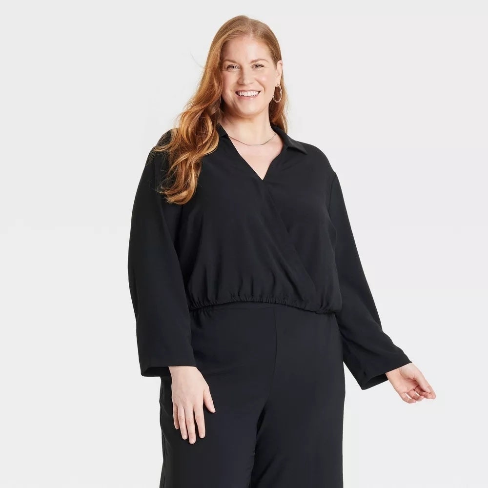 A smiling person wearing a stylish black, long-sleeved wrap top and black pants stands against a plain background