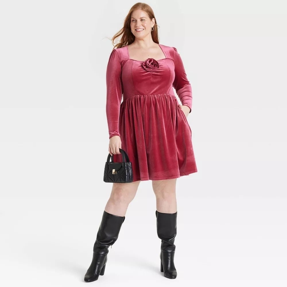 Model in a velvet dress holds a black handbag, wearing knee-high black boots