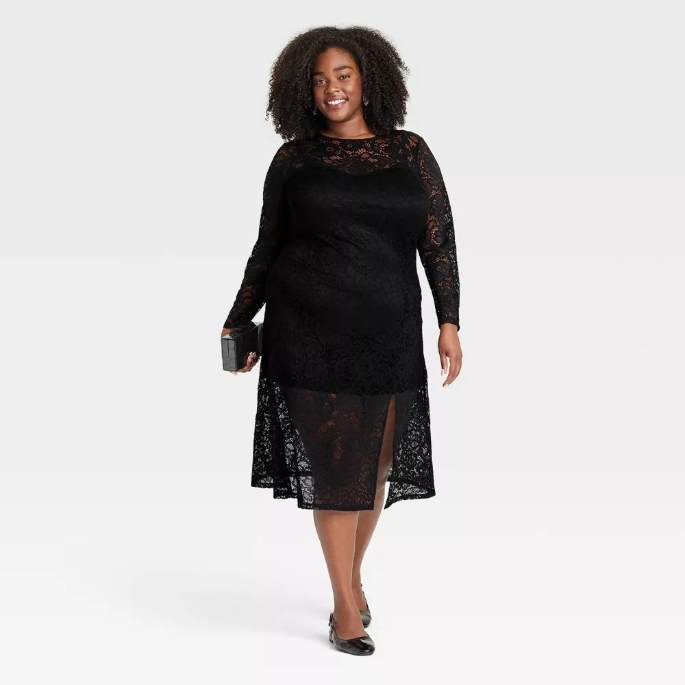 A model in a lace dress with long sleeves and holding a clutch. She is wearing flat shoes