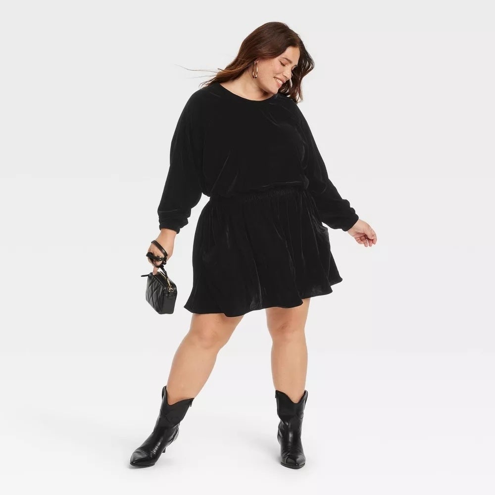 Model in a black long-sleeve dress and boots holds a small purse, smiling and posing confidently