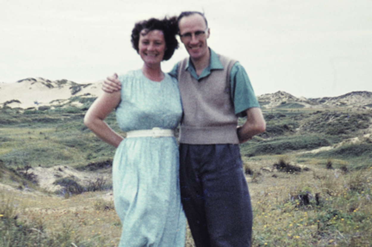 A couple stands together outdoors in a natural setting with sandy hills and grass. The woman wears a dress, and the man wears a sleeveless sweater
