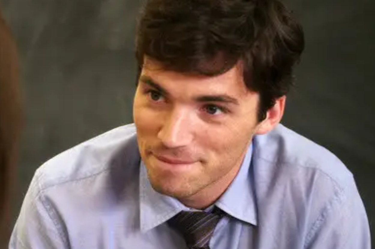 Ezra in a light shirt and tie smiling slightly, looking to the side. The background is blurred