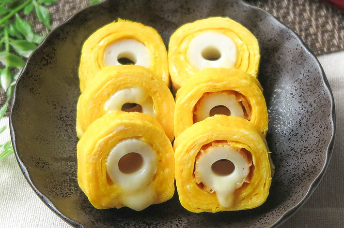 黒い皿に盛られた6つの卵巻きちくわがテーブルに置かれています。写真の背景に箸と植物が見えます。