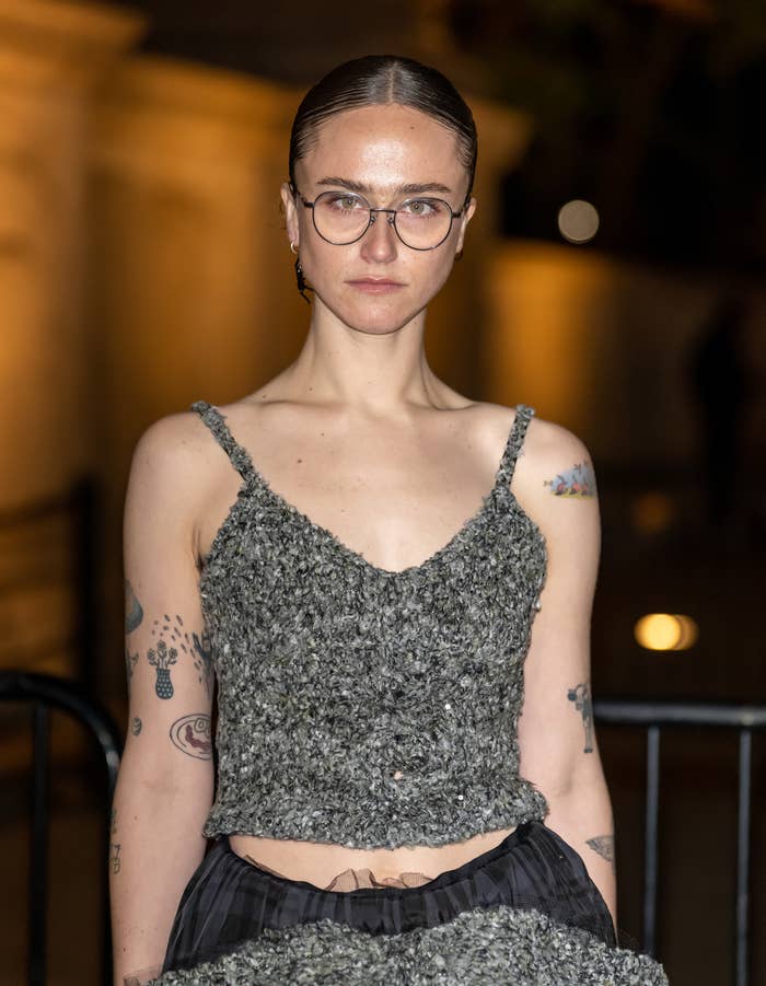 A person with glasses stands on a red carpet wearing a textured sleeveless top and skirt, showcasing tattoos on arms