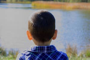 Child with short hair facing a lake, wearing a plaid shirt. The serene outdoor scene suggests a peaceful moment in nature