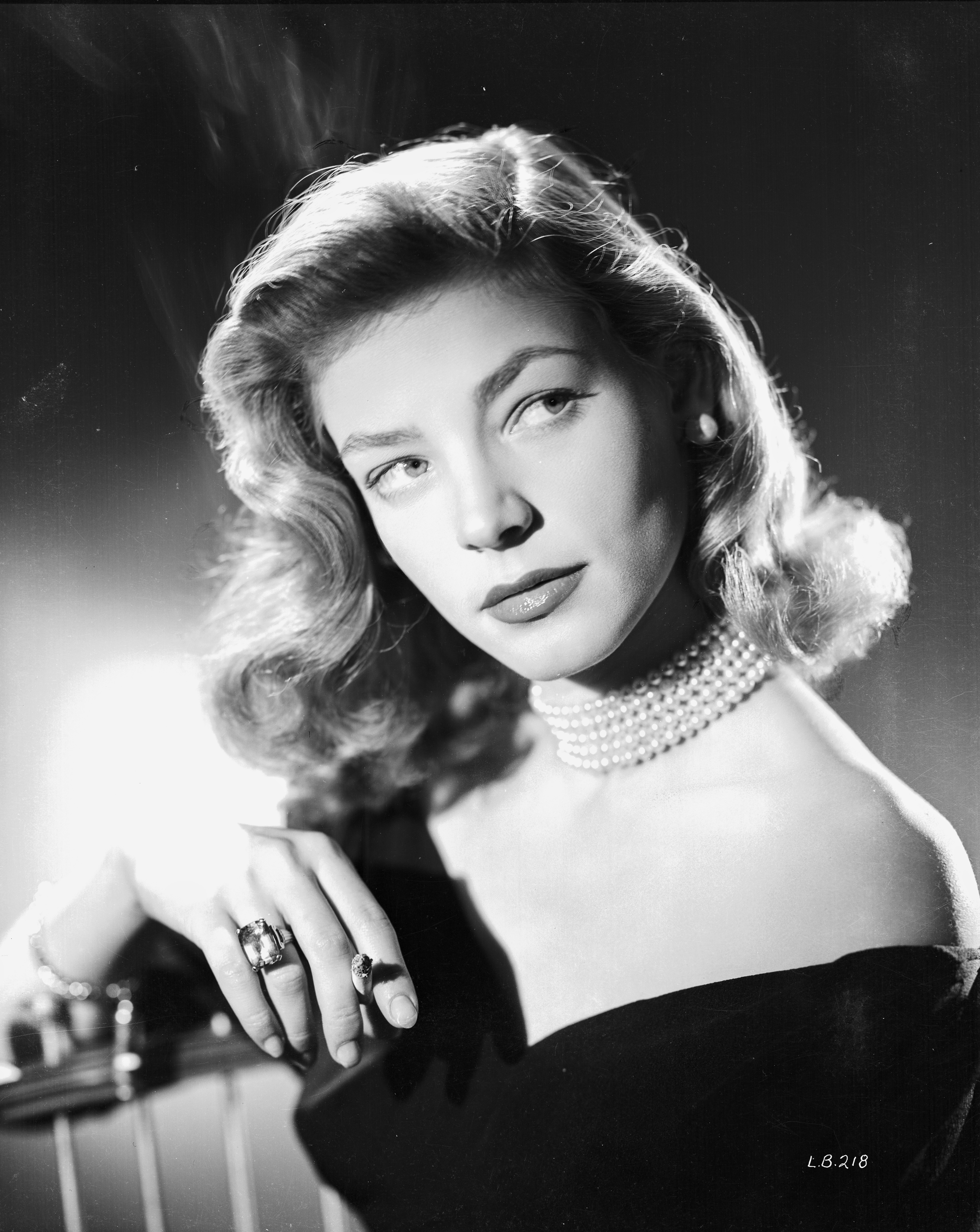 Lauren Bacall, smoking a cigarette, poses for a close-up wearing an off-shoulder dress and multiple pieces of jewelry