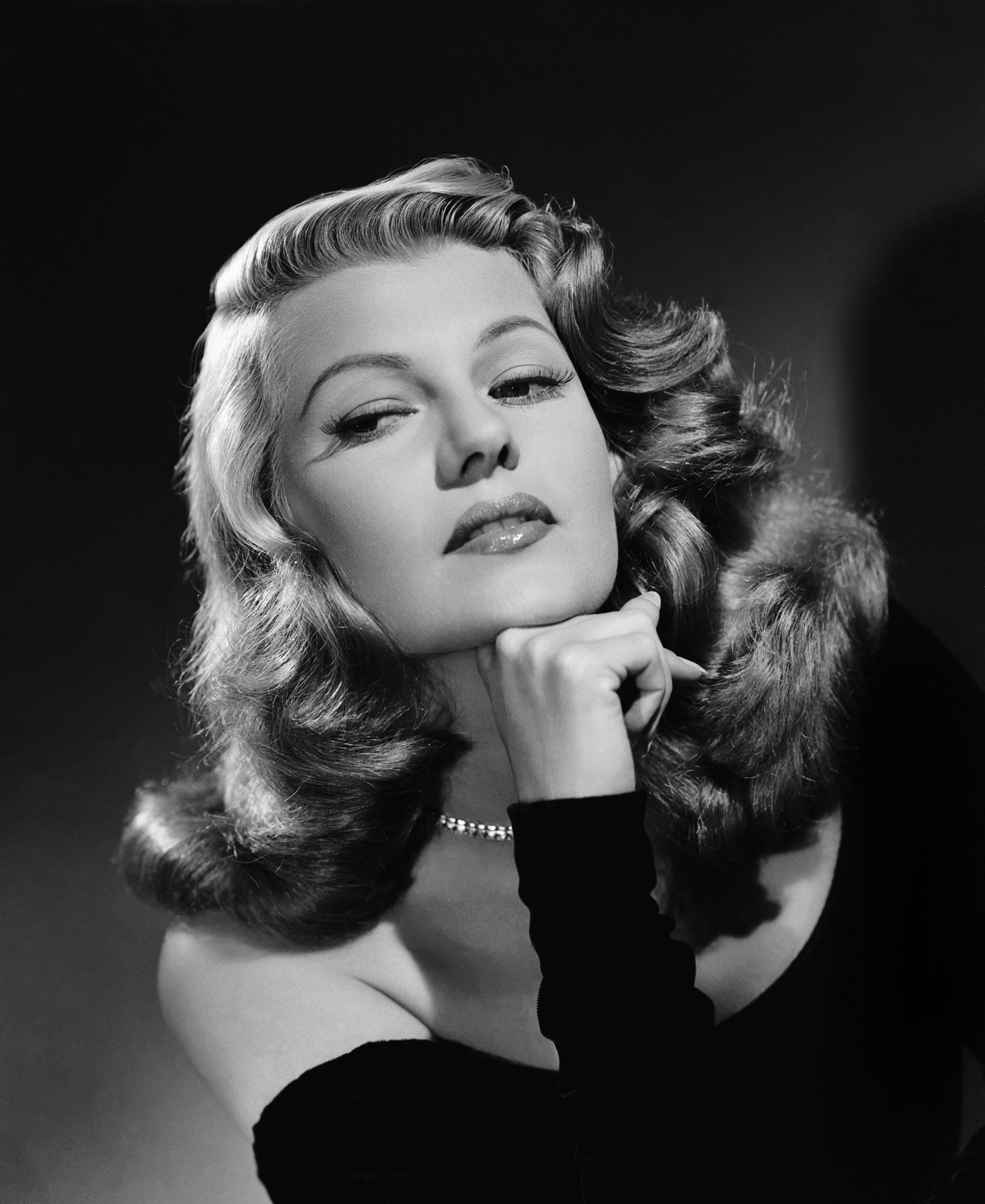 Rita Hayworth wearing a dress and necklace poses with her chin resting on her hand in a vintage glamor shot