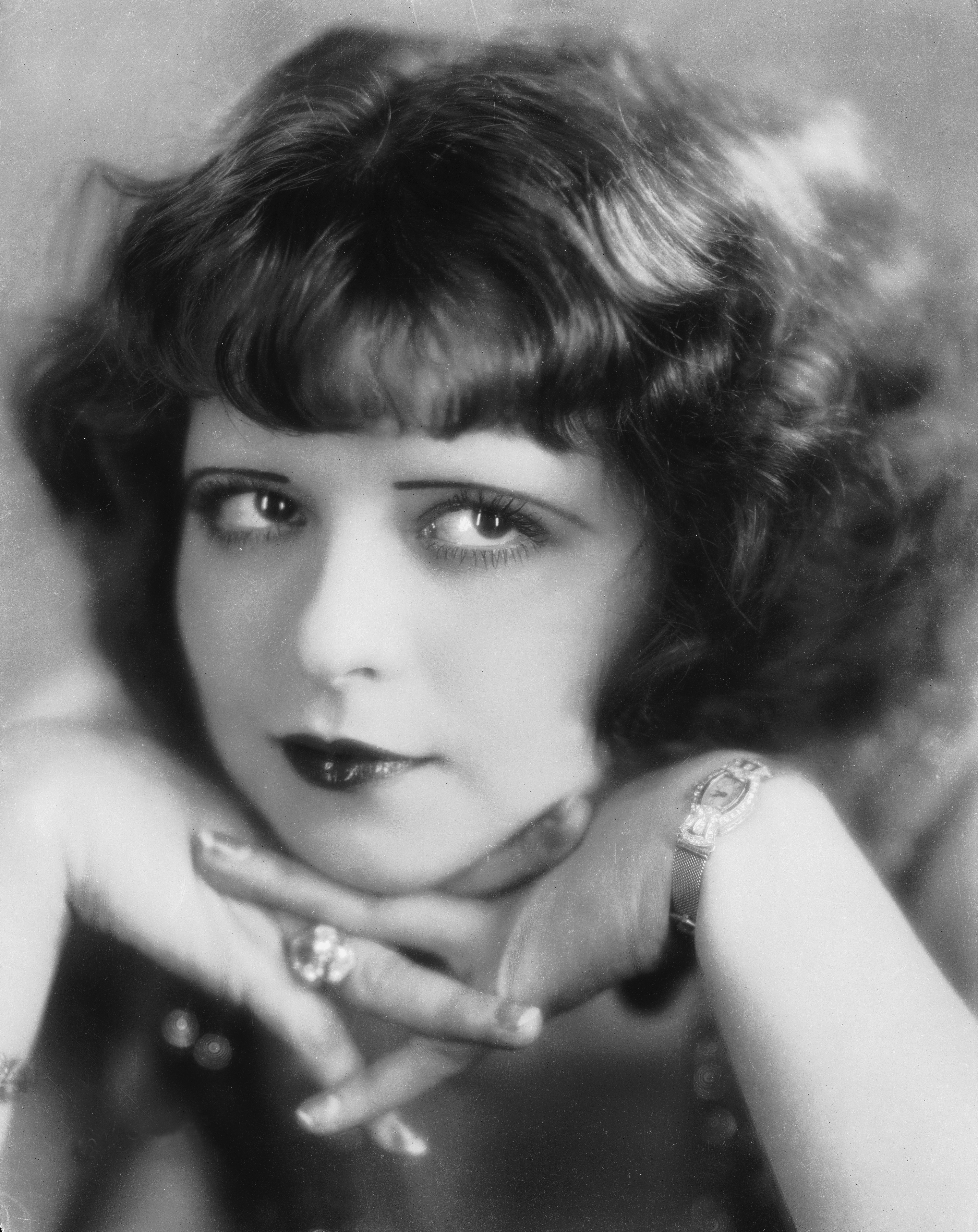 A vintage-style portrait of a woman with curled hair, wearing lipstick and jewelry, resting her face on her hands