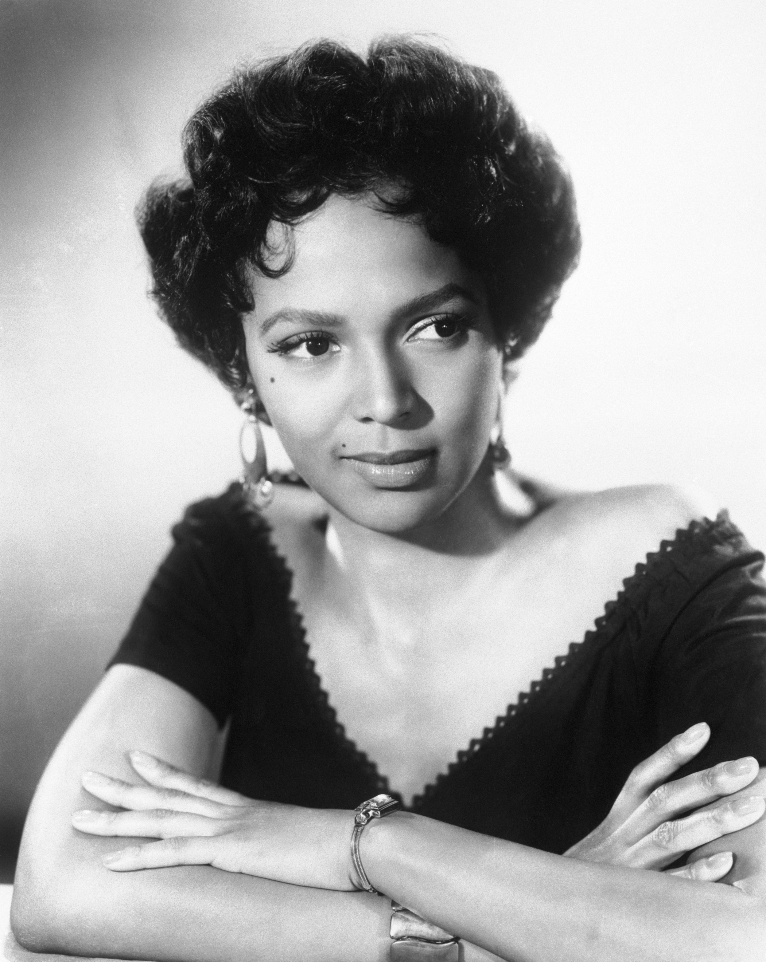 A woman with a classic hairstyle and elegant dress poses with folded arms, wearing hoop earrings and a bracelet, in a black and white portrait