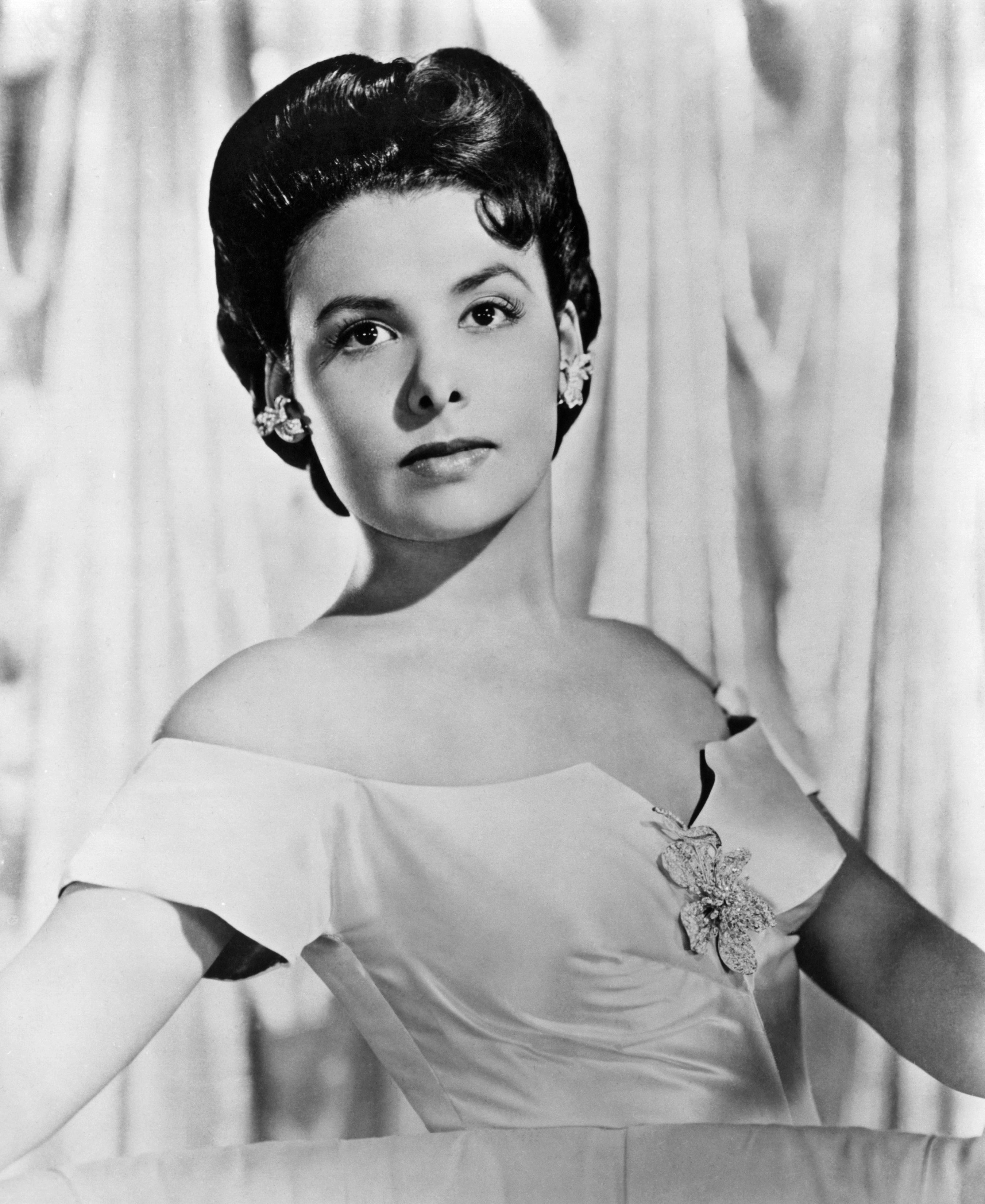 A poised woman in an elegant off-the-shoulder gown, adorned with a floral brooch. Her hair is styled softly, and she wears statement earrings