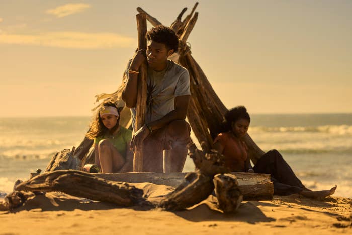 Three people sit on a beach near a driftwood structure, looking pensive. The scene appears serene and introspective with the ocean in the background