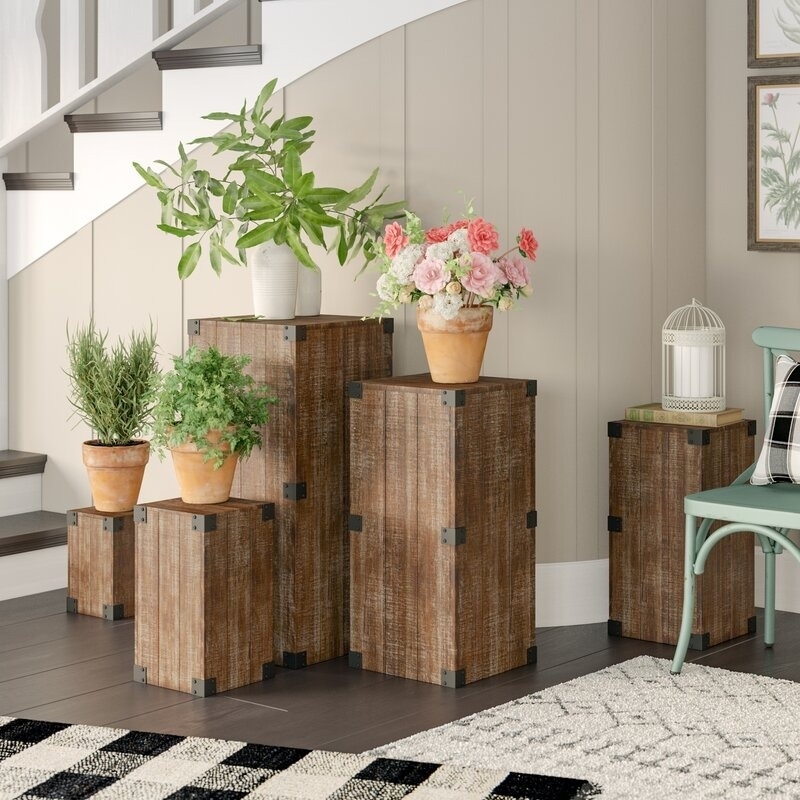 Wooden pedestals display various potted plants and flowers in a cozy indoor setting near a staircase, enhancing the rustic decor