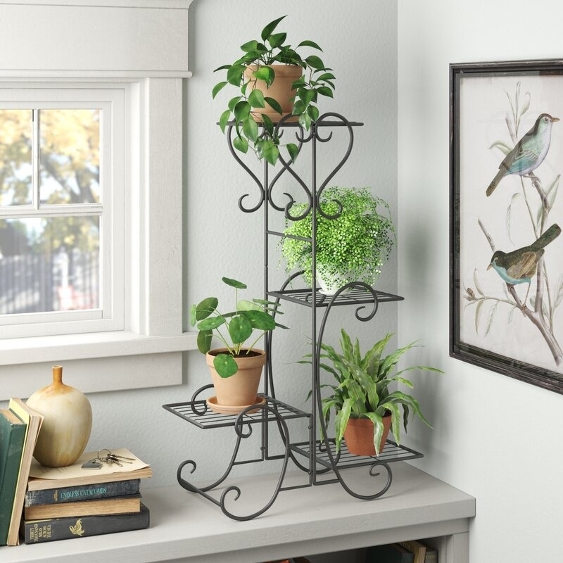 Indoor plant stand with four potted plants on a table by a window. A bird-themed wall art hangs nearby. Books and a decorative gourd are displayed beside it