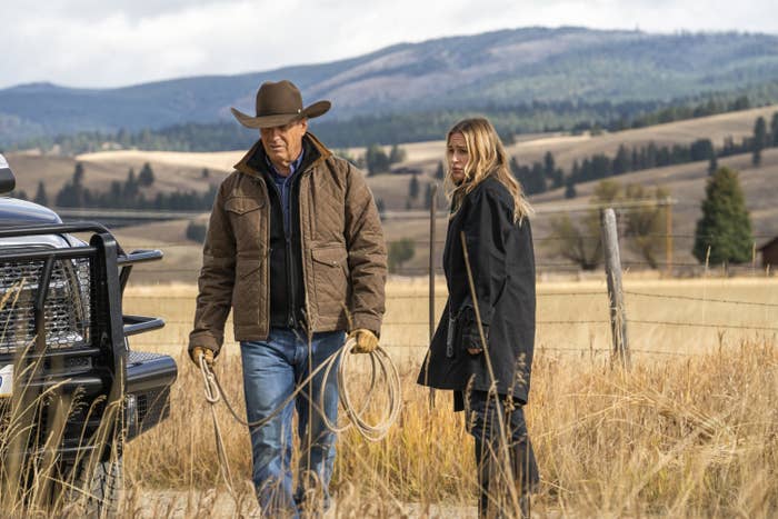 Two people stand in a rural field next to a truck. One wears a cowboy hat and jacket, holding a rope. The other wears a long coat. Mountains are in the background