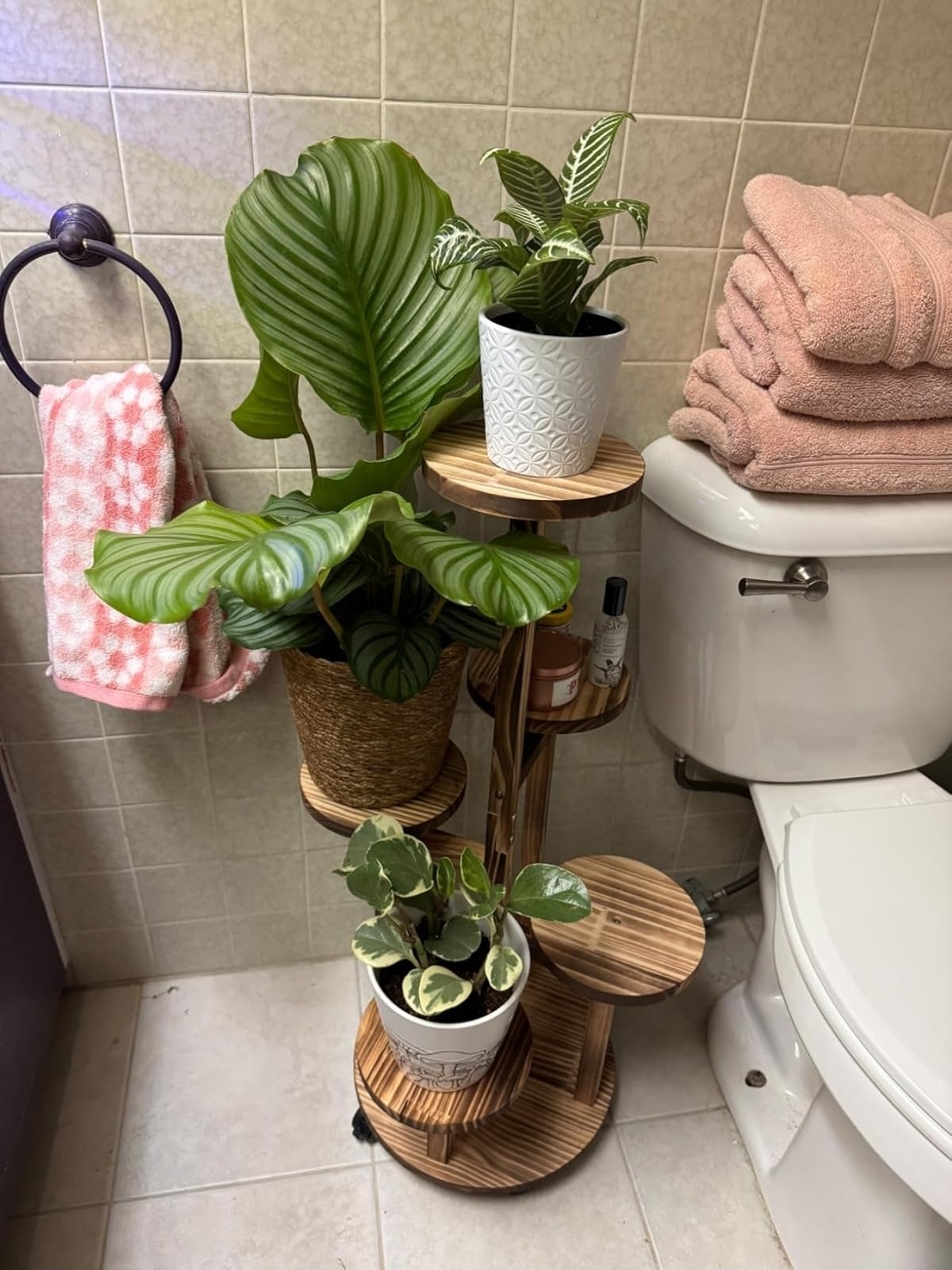 Bathroom decor with potted plants on a wooden stand next to a toilet, pink towels, and a patterned hand towel