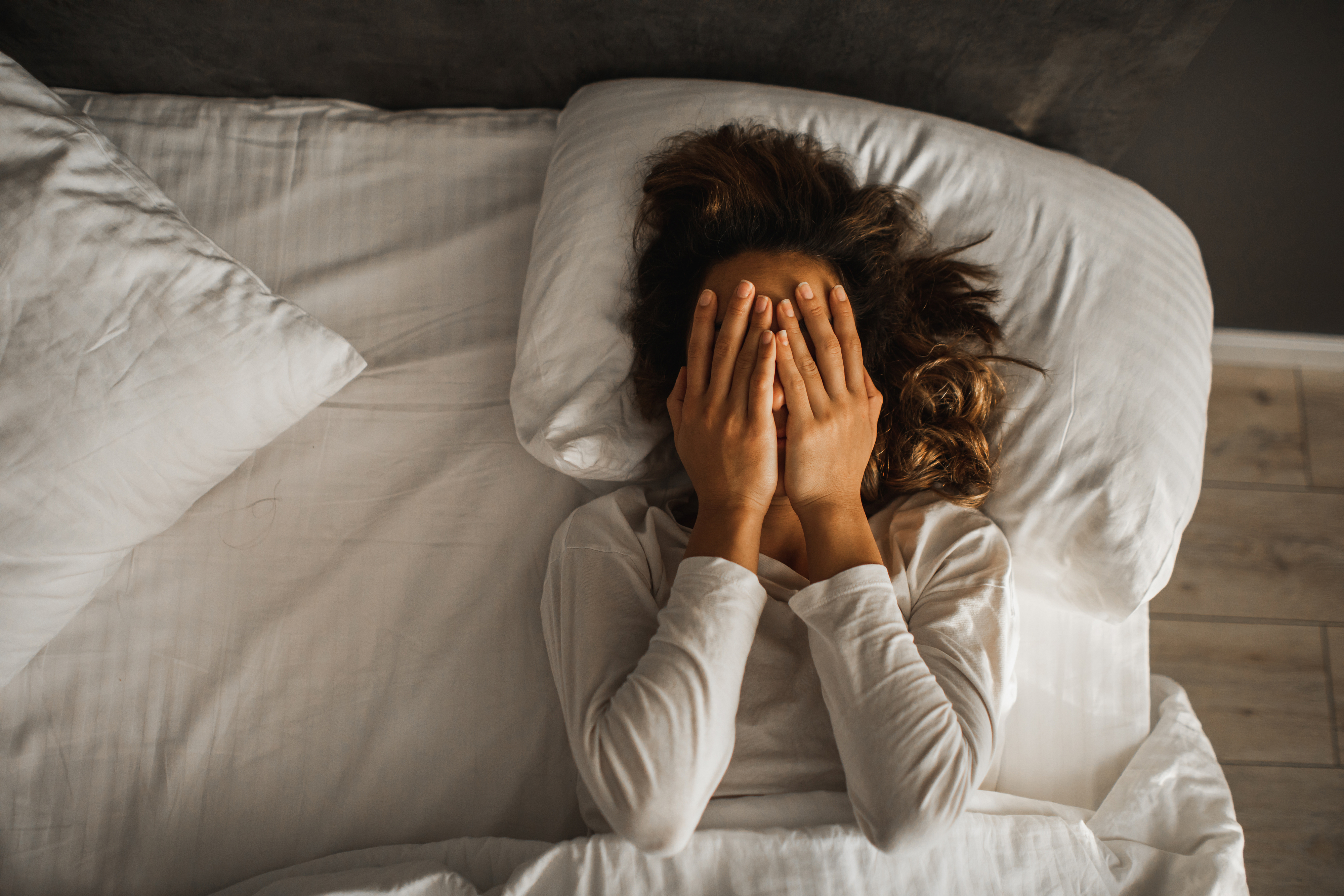 Person lying in bed with hands covering face, wearing a long-sleeve shirt, appearing to express stress or exhaustion