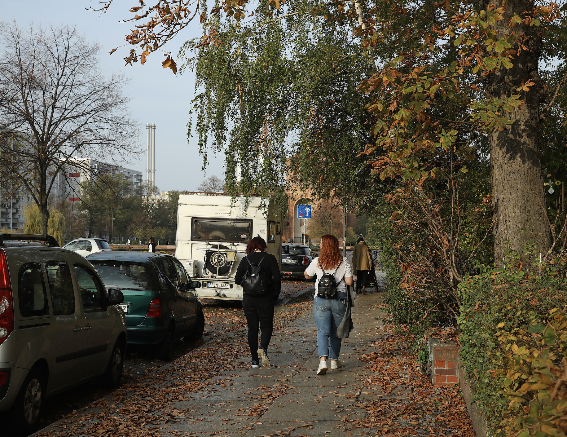 Two people walk down a leafy sidewalk in an urban area, carrying backpacks, with parked cars and trees nearby