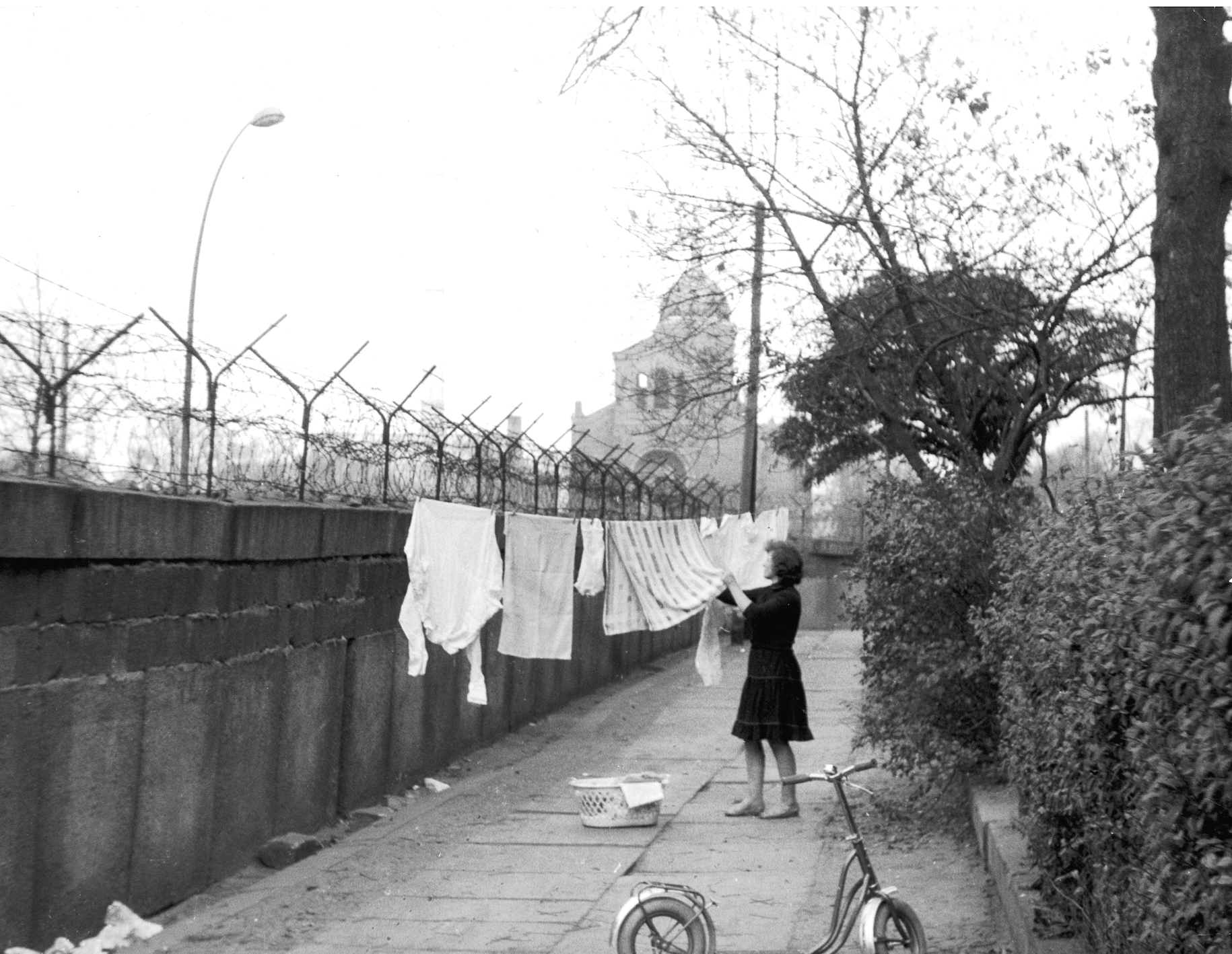 A woman hangs laundry on a line beside a concrete wall topped with barbed wire, a church visible in the background. A bike lies nearby