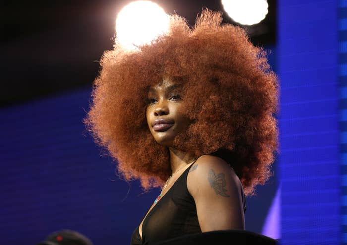 SZA with voluminous hair and a tattoo visible on her arm stands against a lit backdrop, wearing a sleeveless top