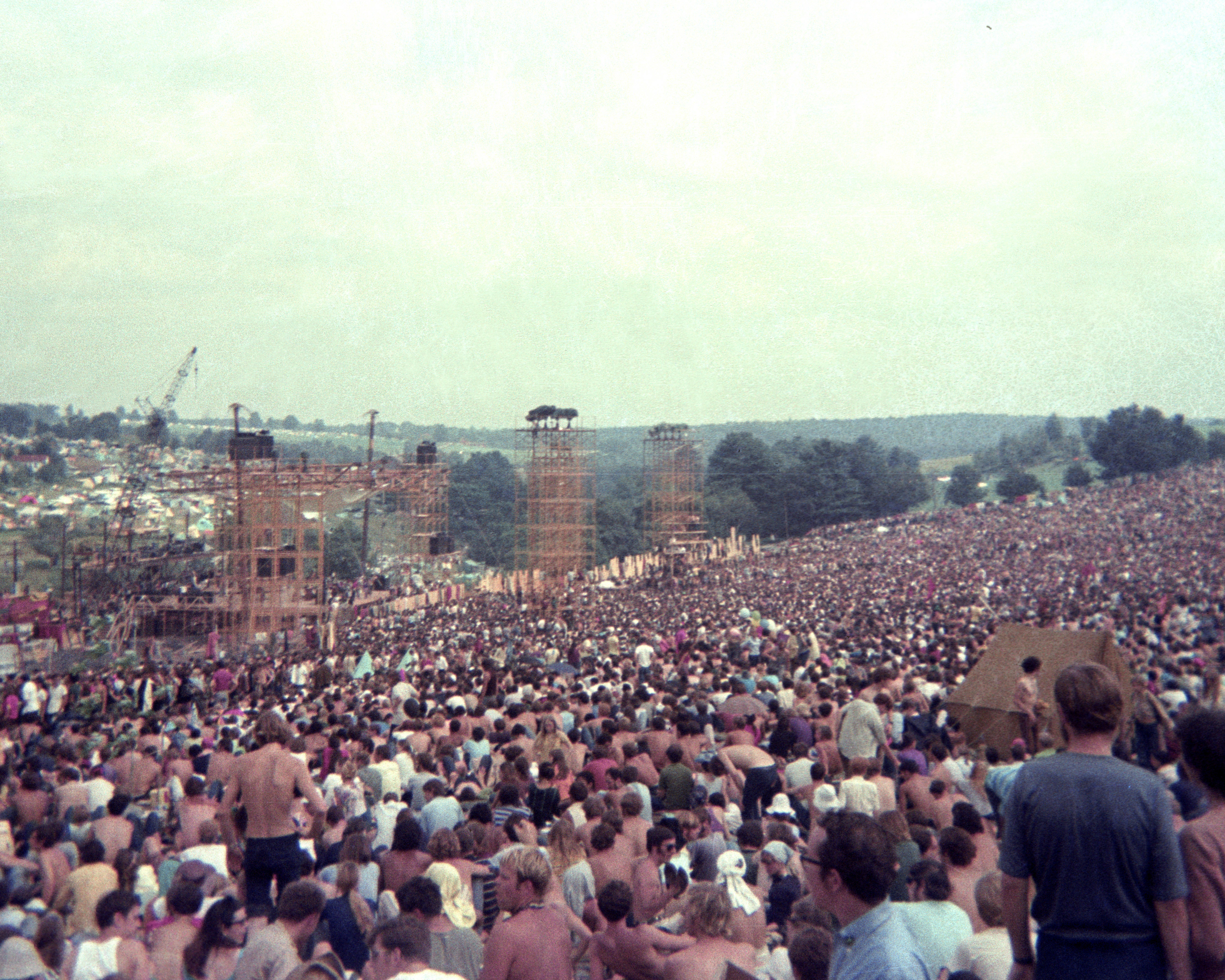 A massive crowd gathers outdoors at a music festival, with a stage and scaffolding visible in the background under a cloudy sky