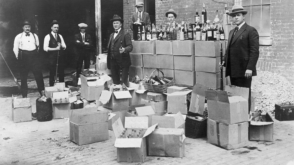Historical photo of Prohibition-era law enforcement officers with confiscated alcohol bottles and boxes, standing outside a brick building