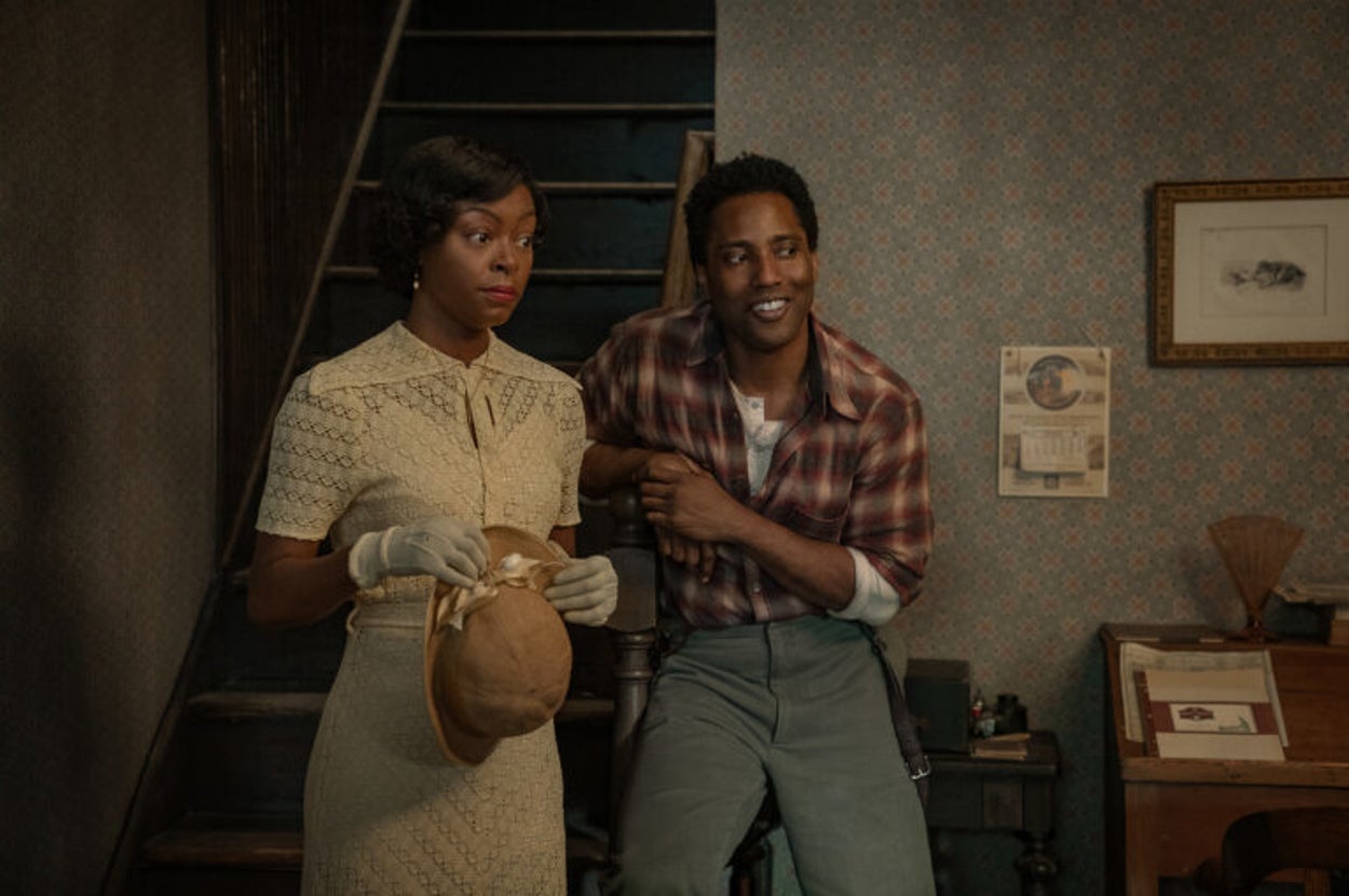 Two people in vintage clothing; a woman in a lace dress with gloves and hat, and a man in a plaid shirt and slacks, standing in a retro interior