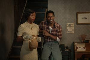 Two people in vintage clothing; a woman in a lace dress with gloves and hat, and a man in a plaid shirt and slacks, standing in a retro interior