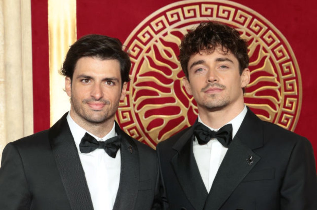 Two men in tuxedos posing on a red carpet with an intricate patterned backdrop