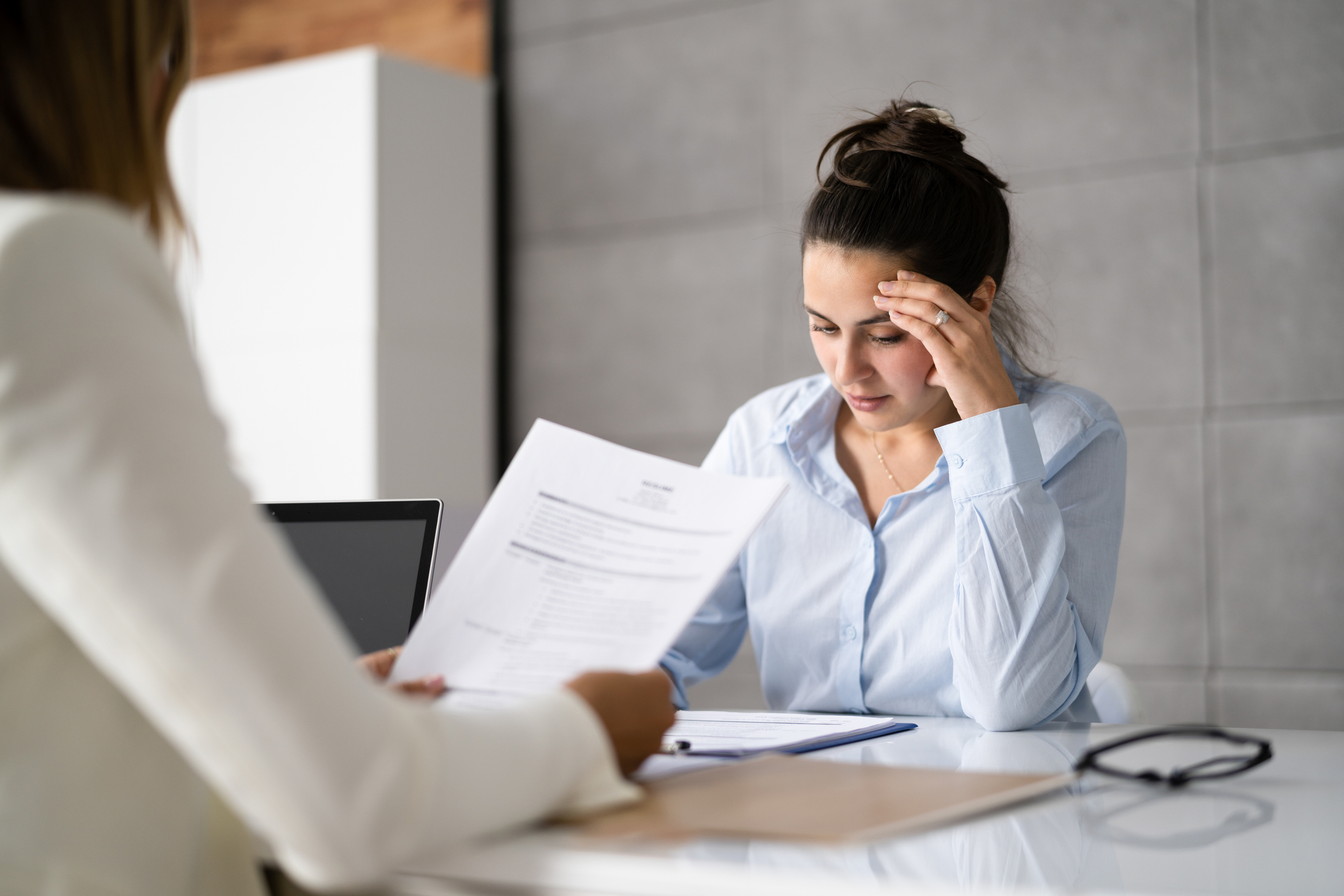 Two people at a desk; one is holding papers, and the other looks thoughtful, touching their forehead in concentration