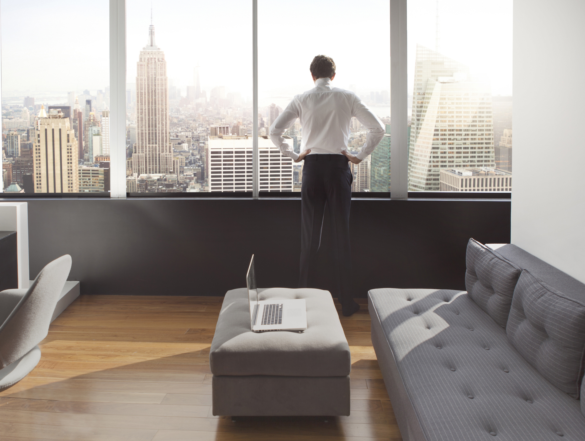 Person stands at a window overlooking a city skyline, hands on hips. A laptop rests on an ottoman in a modern living room