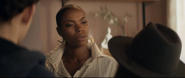 Sasheer Zamata in a TV scene wearing a checkered shirt and large hoop earrings, engaging in conversation with two others