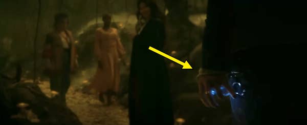 Scene from a TV show with figures in a forest; a person in a cloak stands in the foreground holding a glowing object