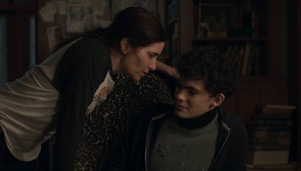 A woman leans toward a seated young man in a cozy, book-filled room, engaging in an intense conversation