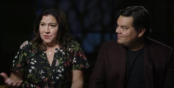 Two people seated, one gesturing while speaking. The woman wears a floral dress, the man a dark jacket. Appears to be an interview setting