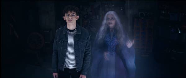Young man in casual clothes stands with a ghostly, elderly woman in a dimly lit room, suggesting a supernatural or mysterious TV/movie scene