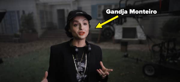 Person in a casual outfit with a hat, wearing a headset, speaks energetically on a film set outdoors
