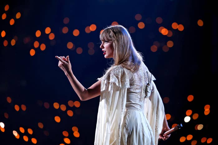 Performer on stage in a flowing, intricate dress, gesturing with one hand, surrounded by blurred lights in the background